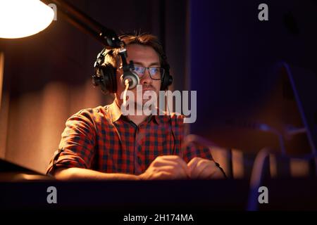 From below focused young male in checkered shirt and eyeglasses using computer and speaking in mic while recording podcast in dark studio Stock Photo