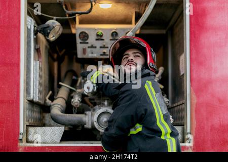 Worker wearing protective uniform and red hardhat operating with pipeline located inside fire truck Stock Photo