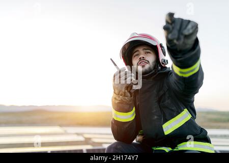 Positive young bearded male firefighter wearing helmet and protective worn gloves and clothes while holding walkie talkie in raised hand and peacefull Stock Photo