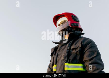 Unrecognizable firefighter wearing protective hardhat and special uniform with yellow reflective patches on background of clear sky in daytime Stock Photo