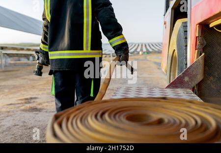 Crop anonymous person in protective uniform carrying end of fire hose while standing next to fire engine with field of solar panels on blurred backgro Stock Photo