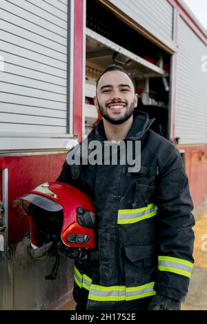 Cheerful bearded young firefighter standing near fire truck and holding red hardhat in hand while smiling and looking at camera Stock Photo