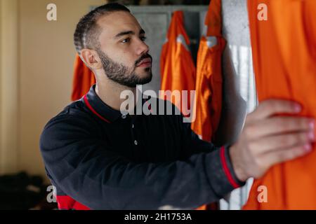 Young handsome bearded ethnic male opening locker and preparing to put on orange uniform on fire station Stock Photo