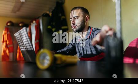Concentrated young bearded ethnic guy wearing shirt sitting at table with laptop and transmitter looking at screen with yellow flashlight in foregroun Stock Photo