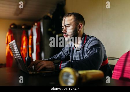 Concentrated young bearded ethnic guy wearing shirt sitting at table with laptop and transmitter looking at screen with yellow flashlight in foregroun Stock Photo
