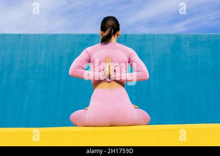 back view of slim female with dark hair wearing sportswear sitting in reverse prayer pose with hands behind back and legs crossed against blue wall 2h1759d