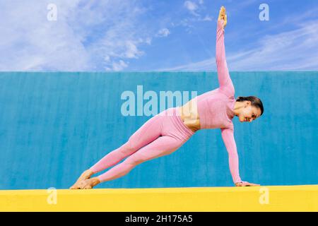 Full length of young slim female concentrating in side plank while practicing yoga Vasishthasana posture on yellow and blue wall Stock Photo