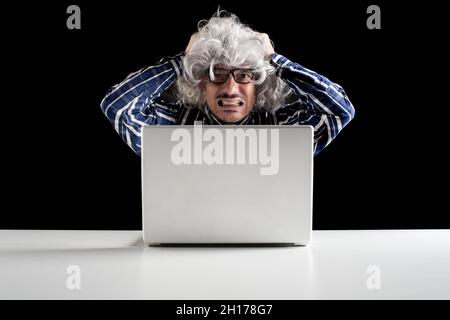 Portrait of a stressed senior man working on a laptop computer sitting at a white table on black background Stock Photo