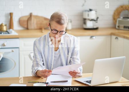 Focused senior woman wearing glasses working with documents while sitting in the modern kitchen Stock Photo