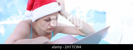 Sad woman in red santa claus hat sitting in front of laptop on sun lounger of swimming pool Stock Photo