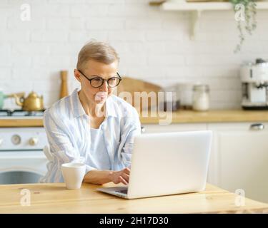 Concentrated senior woman writer typing something on laptop while working online from home Stock Photo