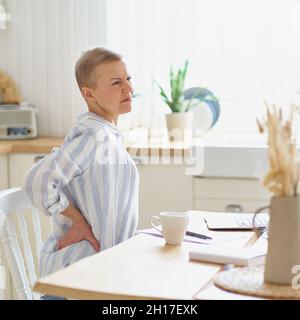 Mature woman massaging her back while working or studying on laptop online at home Stock Photo