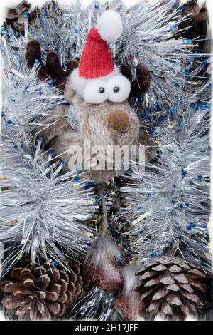 A funny little toy deer with bulging eyes lies in silver tinsel with fir cones. Stock Photo