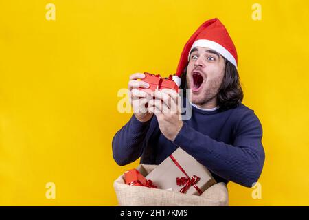 A young man with a Santa Claus hat shows his surprise and joy in picking up a Christmas gift from a sack full of presents. Yellow background for a stu Stock Photo