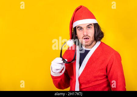 A young surprised Santa! A handsome man dressed like Santa Claus takes off his sunglasses with a surprised expression on his face. Yellow background. Stock Photo