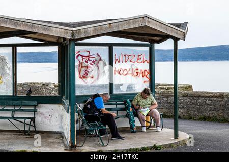 Two people sitting in a run down seaside shelter, with broken windows and graffiti Stock Photo