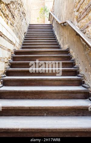 Architectural detail of outdoor wooden old weathered staircase going up with wooden handrail, between two stone bricks walls, in abandoned building Stock Photo