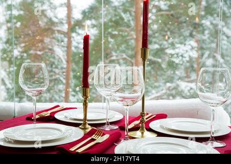 Beautiful table setting with classic white porcelain, golden cutlery and candles. Big window with winter landscape, pine tree forest covered with snow Stock Photo