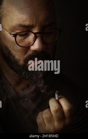A vertical shot of a Hispanic man with glasses and beard smoking and looking at his cigarette Stock Photo