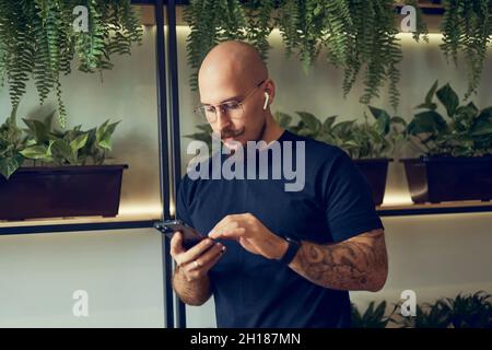 Concentrated businessman uses smartphone with headphones while stands in home office. Distance Job.  Stock Photo