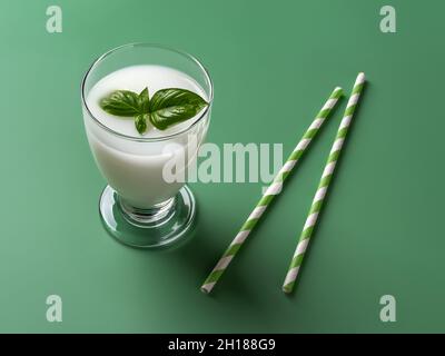 Milk white cocktail with fresh green basil in the footed glass and two stripped paper straws over green background. Disposable cardboard drinking tube Stock Photo
