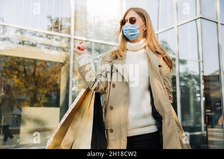 stylish woman in a protective mask against coronavirus, and shopping bags in her hands, in a beige raincoat and sun goggles. against the backdrop of a Stock Photo