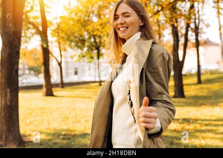 portrait of a beautiful stylish blonde in a beige raincoat posing against the backdrop of an autumn park. leaf fall. yellow leaves on trees. Stock Photo