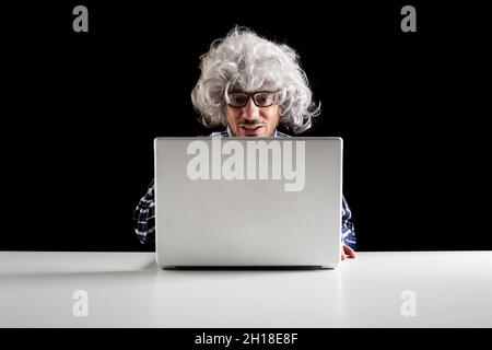 Smiling senior man with gray hair wearing a checkered shirt using laptop sit at the desk. Black background with copy space Stock Photo
