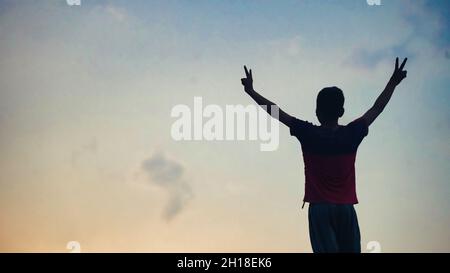 silhouette of boy standing on a roof at sunset. Young adult raising arms on the roof at sunset hands up Stock Photo