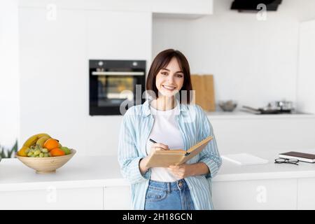Young lady smiling and taking notes, holding notepad and pen, standing in kitchen interior, planning her day Stock Photo