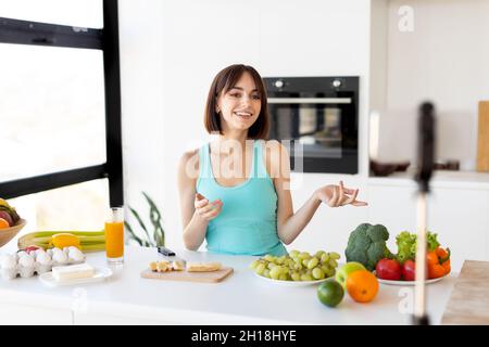 Food blogger concept. Millennial woman recording new video recipe on smartphone, standing in modern kitchen interior Stock Photo