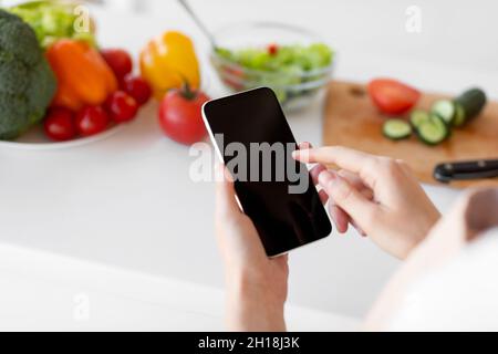 Healthy food and diet planning concept. Young woman using smartphone with blank screen, preparing salad in kitchen Stock Photo