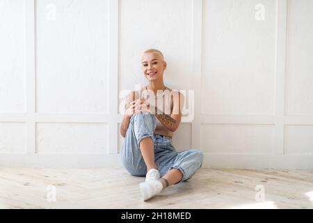 Portrait of young happy caucasian woman with short hair sitting on floor and smiling at camera, leaning on white wall Stock Photo