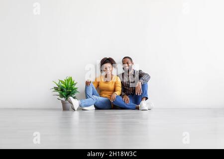 Happy millennial black couple sit on floor with plant in pot on gray wall background in empty room, copy space Stock Photo