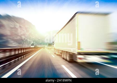 Generic semi trucks speeding on the highway at sunset - Transport industry concept with semitruck containers driving to the mountain pass Stock Photo