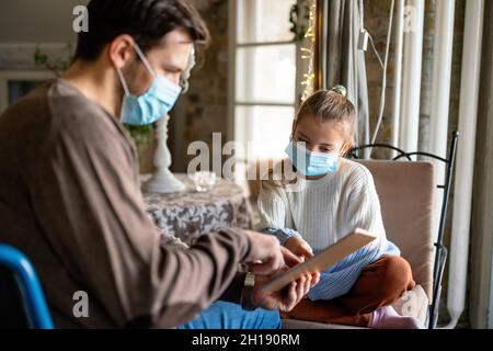 Father with disability in wheelchair using tablet at home with child while wearing masks. Stock Photo