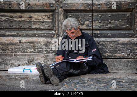 Gray haired man sitting on the ground, leaning against Palazzo San Calisto door and sketching by Piazza di Santa Maria in Trastevere, Rome, Italy Stock Photo