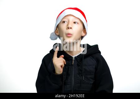 a boy in a red Santa hat who stands on a white background holds his finger to his lips and thinks Stock Photo