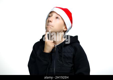 a boy in a red Santa hat who stands on a white background holds his finger to his lips and thinks Stock Photo