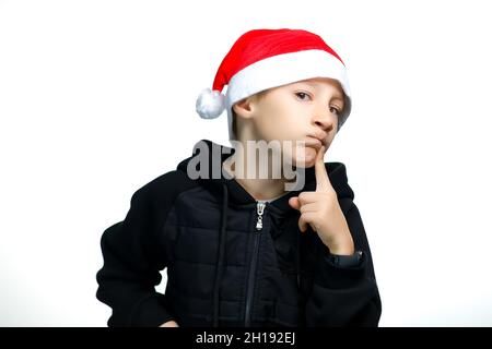 a boy in a red Santa hat who stands on a white background holds his finger to his lips and thinks Stock Photo