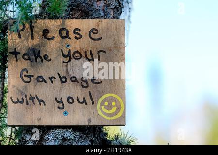 A hand painted sign in the woods asking people to take their garbage when they leave. Sign is attached to a tree. Smiley face drawn on sign. Stock Photo