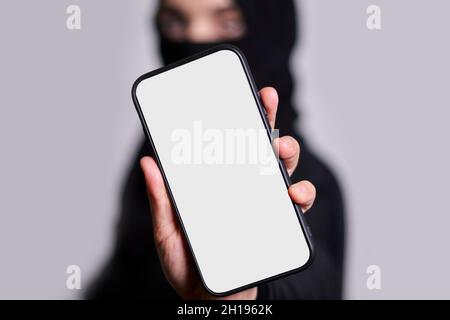 Muslim woman in hijab holds a smartphone with empty screen. Arab woman in traditional clothing holding mobile phone with blank white screen. Copy Stock Photo