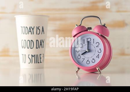 Pink alarm clock with a paper cup of coffee on wooden desk. Minimal time concept. Stock Photo