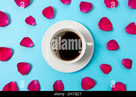 Creative composition of red rose petals and cup of coffee on blue background. Minimal love concept. Stock Photo