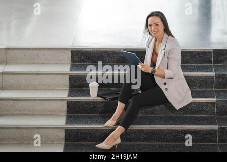 A cheerful Thai young businesswoman sitting on the stairs working on her tablet in the office Stock Photo