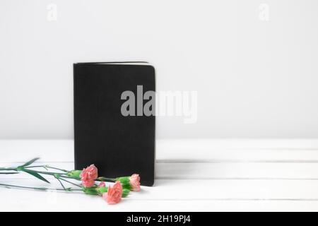 White wooden desk with black empty book or notebook and pink flowers. Front view. Place for text, copy space Stock Photo