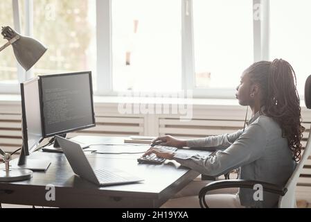 Minimal side view portrait of young African-American woman using computer and writing code while sitting against window in software development office Stock Photo