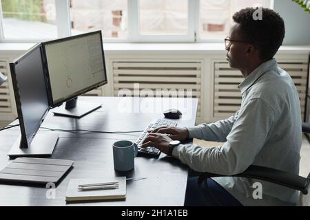 Side view portrait of focused African-American man using computer while sitting at desk in office, software developer concept, copy space Stock Photo