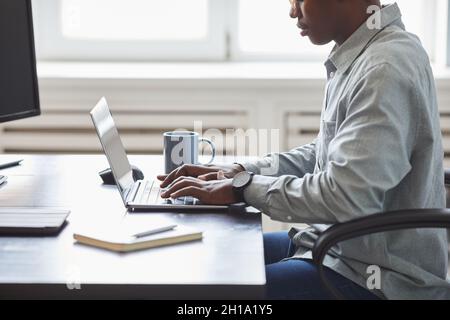 Minimal side view shot of young African-American man using laptop while sitting at desk in office, focus on hands typing at keyboard, software develop Stock Photo