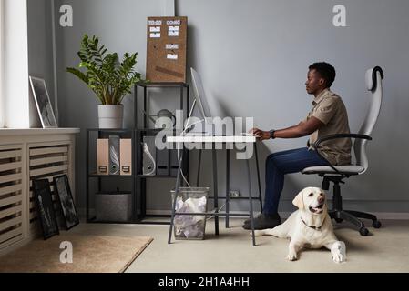 Full length portrait of young African-American man working in office with dog laying on floor, pet friendly workspace, copy space Stock Photo